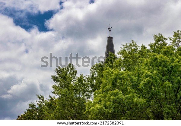 A top of a building\'s steeple rises above\
the tops of trees and into cloudy\
skies.