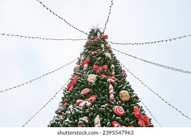 The top of the Christmas tree at the New Year fair is decorated with candies and red garlands. Light bulbs on wires radiate from the top of the tree



