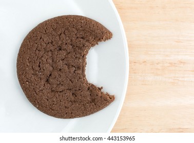 Top Close View Of A Dutch Cocoa Cookie That Has Been Bitten On A White Plate Atop A Wood Table Top.