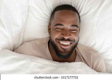 Top close up view laughs handsome african guy lying in bed woke up feel peppy and healthy sleeping enough hours. Under white warm blanket on fresh soft sheets comfortable mattress resting cheerful man