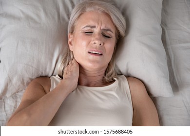 Top Close Up View Middle-aged Woman Lying In Bed In Morning Feels Pain In Neck After Night Sleep, Awaken Having Painful Sudden Ache Or Stiffness, Incorrect Posture During Asleep, Soft Mattress Concept