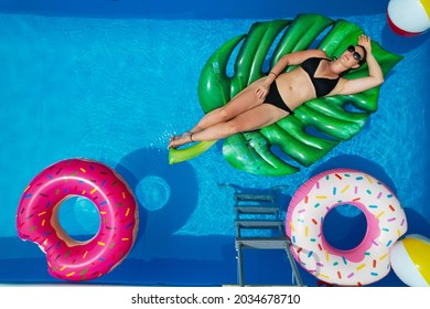 TOP DOWN, AERIAL, COPY SPACE: Beautiful Young Woman Lies On A Big Leaf Floatie In Her Crystal Clear Home Pool. Caucasian Female In A Black Bikini Relaxes In Her Garden Pool On A Sunny Summer Weekend.