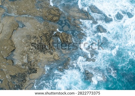 Top down aerial drone view of powerful blue ocean waves splashing against a dangerous rocky coastline