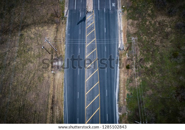 Top down aerial drone view\
Flat looking down from streets. Yellow caution lines and Empty road\
with open fields and green lot on both sides near Round Rock\
Texas