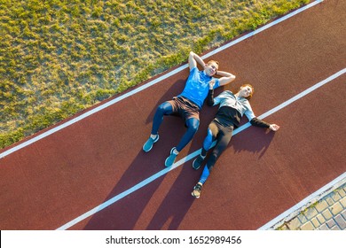 Top Down Aerial View Of Two Young People Sportsman And Sportswoman Laying On Red Rubber Running Track Of A Stadium Field Resting After Jogging Marathon In Summer.