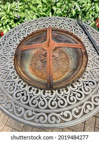 The Top Down, Close Up Image Of A Backyard Fire Pit. It's Currently Empty, Is Covered With A Rusty Metal Lid, And Has A Fire Pick Laying On The Ledge. The Rod Iron Fire Pit Has A Beautiful Design.