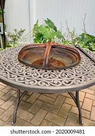 The Top Down, Close Up Image Of A Backyard Fire Pit. It's Currently Empty, Is Covered With A Rusty Metal Lid, And Has A Fire Pick Laying On The Ledge. The Rod Iron Fire Pit Has A Beautiful Design.