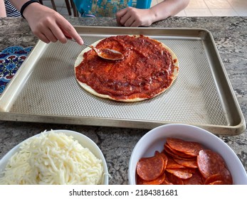 The Top Down, Close Up Image Of A Child Making A Homemade Pizza On A Cauliflower Crust. She's Spooning Sauce And Has Bowls Filled With Toppings.