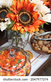The Top Down, Close Up Image Of A Fall Themed Table Setting. A Bouquet Of Orange Sunflowers, A Bowl Filled With Mixed Nuts In The Shell, And A Glass Cookie Jar Filled With Candy Corn Sit In A Tray.