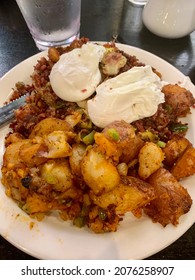 The Top Down, Close Up Image Of A Hearty Breakfast. The Dish Is Overflowing With Corn Beef Hash, Potatoes, And Two Poached Eggs. There Is Also A Coffee Cup And Water On The Table.