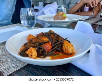The Top Down, Close Up Imagine Of A White Dish With Squid Ink Pasta, Shrimp, And A Tomato Based Sauce. This Delicious Meal Is On A Table In An Outdoor Dining Restaurant.