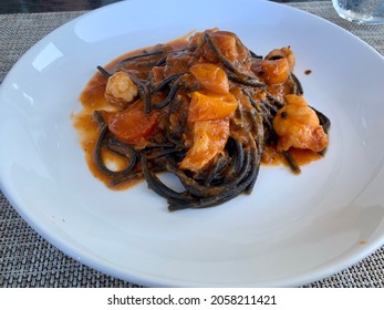 The Top Down, Close Up Imagine Of A White Dish With Squid Ink Pasta, Shrimp, And A Tomato Based Sauce. This Delicious Meal Is On A Table In An Outdoor Dining Restaurant.