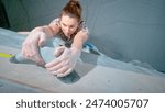 TOP DOWN, CLOSE UP, DOF: Female climber hangs from a rough grey cup hold after topping an artificial boulder. Detailed shot of woman