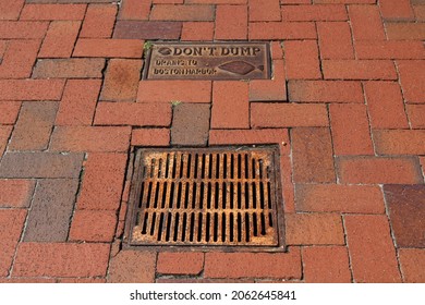 The top down, close up view of a drainage system in the street. The sidewalk is paved with bricks and has a sign that reads "Don't Dump Drains to Boston Harbor" to warn pedestrians before polluting.