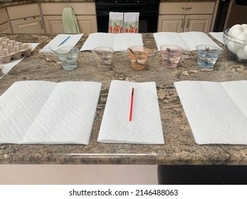 The Top Down, Close Up View Of A Kitchen Counter Set Up For Kids To Paint Easter Eggs. There Are Paper Towels, Paint Brushes, Empty Egg Cartons, Hard Boiled Eggs, And Dying Stations.