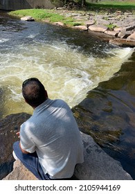 The Top Down, Close Up View Of A Man Hiking Across Boulders And Watching The Water Flow. It Looks Like A Waterfall Or Rapids.