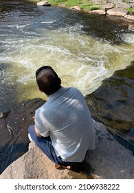 The Top Down, Close Up View Of A Man Hiking Across Boulders And Watching The Water Flow. It Looks Like A Waterfall Or Rapids.