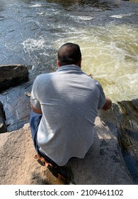 The Top Down, Close Up View Of A Man Hiking Across Boulders And Watching The Water Flow. It Looks Like A Waterfall Or Rapids.