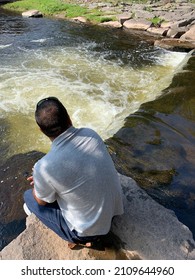 The Top Down, Close Up View Of A Man Hiking Across Boulders And Watching The Water Flow. It Looks Like A Waterfall Or Rapids.