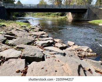The top down, close up view of a small waterfall foaming as the freshwater falls down into the stream below. The boulders are covered in fallen leaves and the water flows under an overpass.