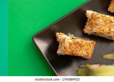 Top Down Close Up View Of Sushi Rolls With Sesame Seeds And Cucumber On A Black Square Plate. Flat On A Bright Green And Light Green Background