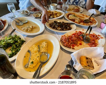 The Top Down, Close Up View Of A Table Filled With Food And Wine Being Served Family Style. There Are People Around The Table And Hands Reaching For Different Dishes.