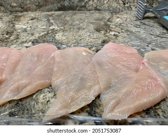 The Top Down, Close Up View Of Thin Sliced Chicken Cutlets That Are Laying On Top Of A Sheet Of Plastic Wrap.