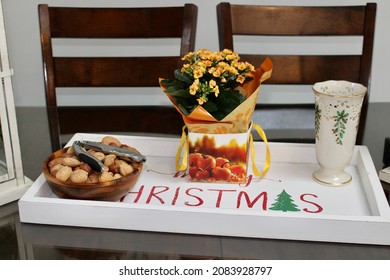 The Top Down, Close Up View Of A Tray Sitting On Top Of A Kitchen Table. The Tray Reads MERRY CHRISTMAS And Is Holding A Nut Bowl, An Orange Primrose Plant, And An Empty Vase With Holly On It.