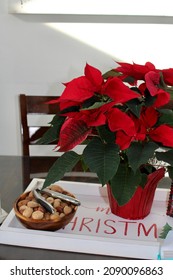 The Top Down, Close Up View Of A Tray Sitting On Top Of A Kitchen Table. The Tray Reads MERRY CHRISTMAS And Is Holding A Nut Bowl, A Bright Red Poinsettia Plant, And A Lantern Filled With Green Balls.