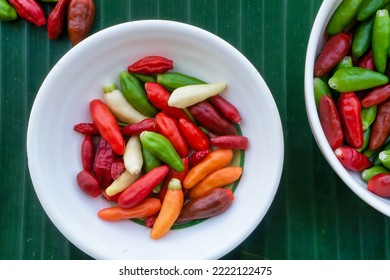 Top down, closeup view of small red, green, orange and creamy white chilli peppers in white dishes on a green banana leaf background. Philippines.