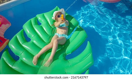 TOP DOWN: Cute Girl Lying On A Floatie Splashing With Water Gun Towards Camera. Cheerful Little Girl Floating In Backyard Swimming Pool And Having Fun With Water Blaster Spraying In Camera Direction.