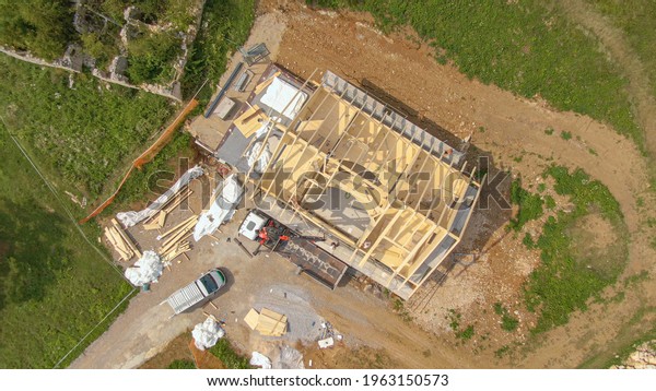 TOP DOWN: Drone point of view of workers building\
the roof of a modern cross-laminated timber house under\
construction in the countryside. Boom lifts wooden beams to the\
roof of prefabricated\
house.