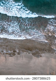Top down drone view captures sea waves with white foam rolling onto a sandy and rocky shore with textured water.