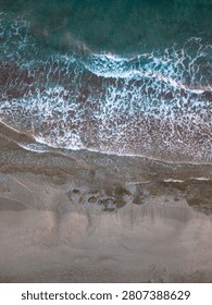 Top down drone view captures sea waves with white foam rolling onto a sandy and rocky shore with textured water.