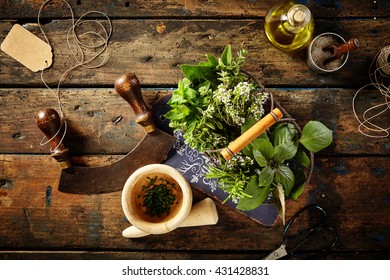 Top Down First Person Perspective View On Various Herbs, Oil, Pestle, Blank Tag And Mezzaluna Over Old Wooden Table