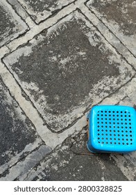 Top down high angle view of a weathered, textured square concrete paving stone walkway with a vibrant blue plastic perforated utility stool on the ground