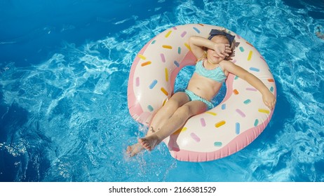 TOP DOWN: Little Girl In Pool Splashing Water On A Floatie Donut Hiding Her Face. Cute Girl Covering Her Face With Hand While Floating And Playing In Backyard Pool. Fun Activity For Summer Holidays.