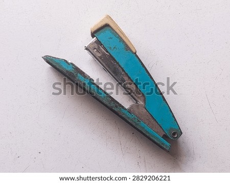 A top down photograph capturing an old, weathered blue metal stapler resting open on a off white, scratched surface. The stapler features chipping blue paint, rusty edges, and a cream-colored plastic cap on its top handle, showcasing significant wear and age. Its metal mechanism inside shows discoloration and patina from long term use. The flat background emphasizes the rustic, vintage character of this classic stationery item.