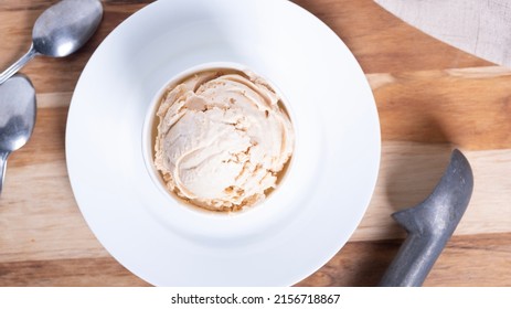 Top Down Of Scoop Of Peanut Butter Or Caramel Ice Cream In A White Bowl With Spoon And Ice Cream Scooper Next To It On A Wood Grain Table