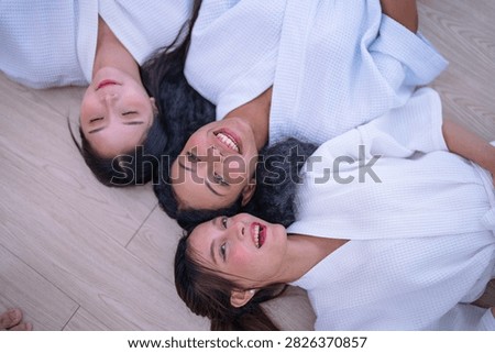 Top down shot of three young female friends resting side by side on a wooden floor in white spa robes as the Asian friend drifts into a quiet moment of rest in a bright white spa room.