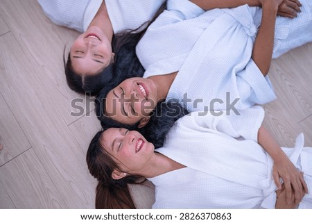 Top down shot of three young female friends resting side by side on a wooden floor in white spa robes as the Asian friend closes her eyes and lifts the corners of her mouth.