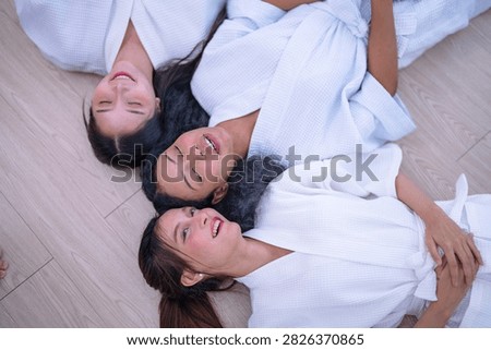 Top down shot of three young female friends resting side by side on a wooden floor in white spa robes as the woman with curly hair rests one arm above her head and relaxes.