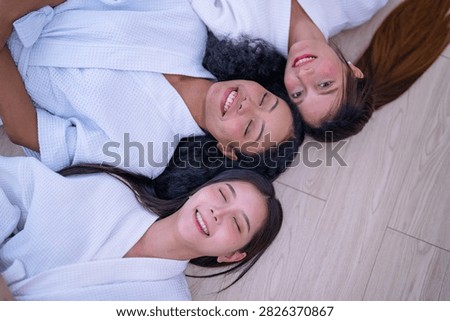 Top down shot of three young female friends resting side by side on a wooden floor in white spa robes as the woman with curly hair holds a peaceful expression without moving.