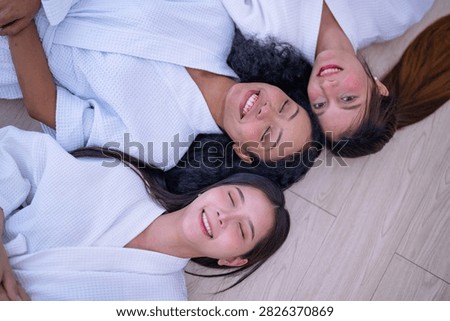 Top down shot of three young female friends resting side by side on a wooden floor in white spa robes as the friend with long brown hair turns her cheek against the warm floor.