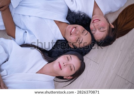 Top down shot of three young female friends resting side by side on a wooden floor in white spa robes as the Asian friend lets a slow smile spread while her eyes stay shut.