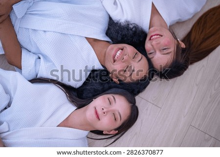 Top down shot of three young female friends resting side by side on a wooden floor in white spa robes as the Asian friend relaxes her whole face into an easy calm in a bright white spa room.