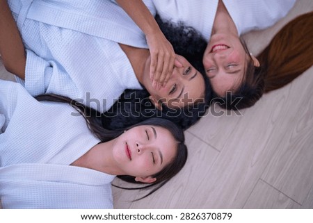 Top down shot of three young female friends resting side by side on a wooden floor in white spa robes as the Asian friend settles deeper into the floor with a soft sigh.