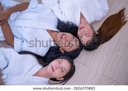 Top down shot of three young female friends resting side by side on a wooden floor in white spa robes as the woman with curly hair keeps her eyes closed while the others start to smile.