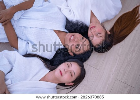 Top down shot of three young female friends resting side by side on a wooden floor in white spa robes as the Asian friend keeps her eyes closed while the others start to smile.