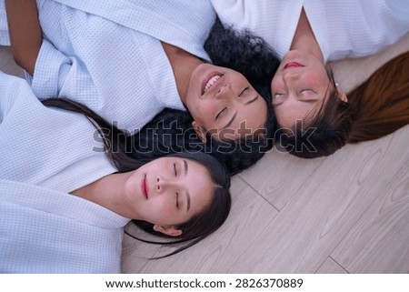 Top down shot of three young female friends resting side by side on a wooden floor in white spa robes as the friend with long brown hair smiles faintly with her eyes closed.